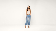 A woman stands against a plain white background, wearing a sleeveless beige top, light blue wide-leg jeans, a belt, and tan high heels. She has long brown hair and a neutral expression, with one hand in her pocket.