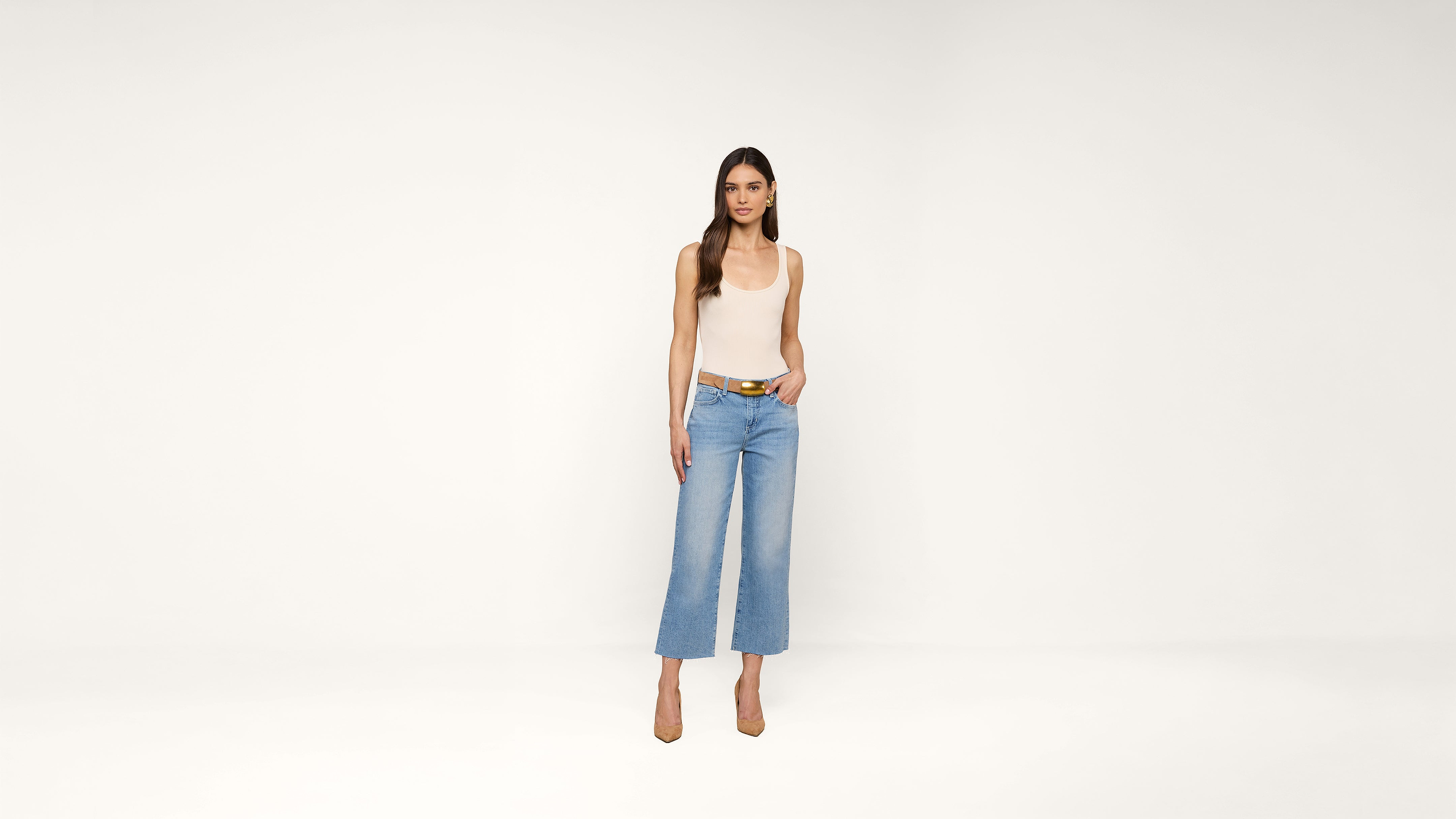 A woman stands against a plain white background, wearing a sleeveless beige top, light blue wide-leg jeans, a belt, and tan high heels. She has long brown hair and a neutral expression, with one hand in her pocket.