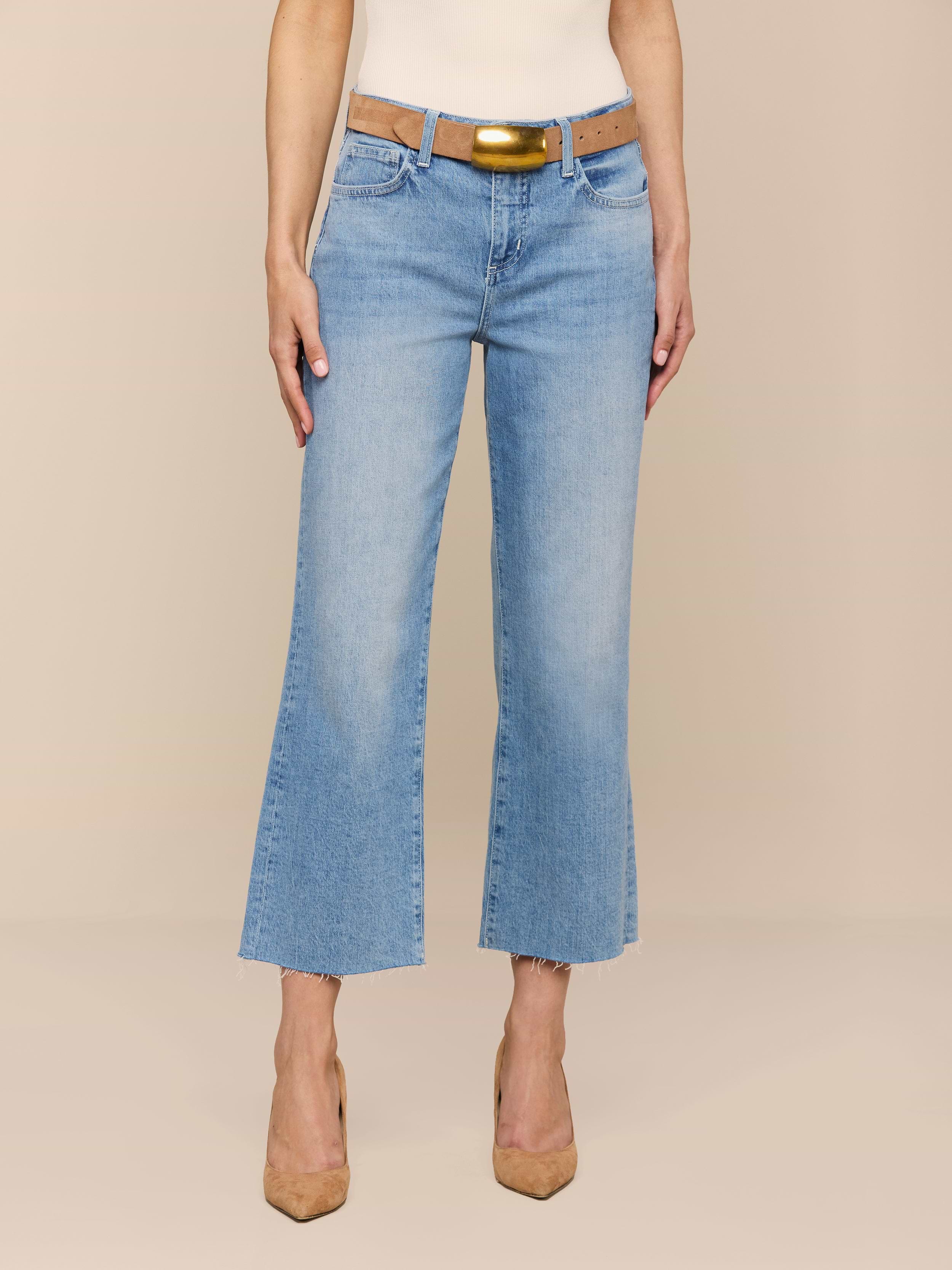Eine Person, die die Wanda Cropped Wide-Leg Jean in hellblauer Waschung mit ausgefransten Säumen trägt, gepaart mit einem hellbraunen Gürtel mit goldener Schnalle, einem weißen Top und hellbraunen spitzen Absätzen, steht vor einem beigen Hintergrund.