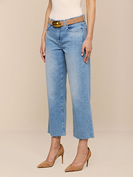Eine Person modelliert die Wanda Cropped Wide-Leg Jean in hellblauer Waschung mit offenem Saum, gestylt mit einem hellbraunen Gürtel, einem weißen Oberteil und hellbraunen hochhackigen Schuhen vor einem schlichten beigen Hintergrund.