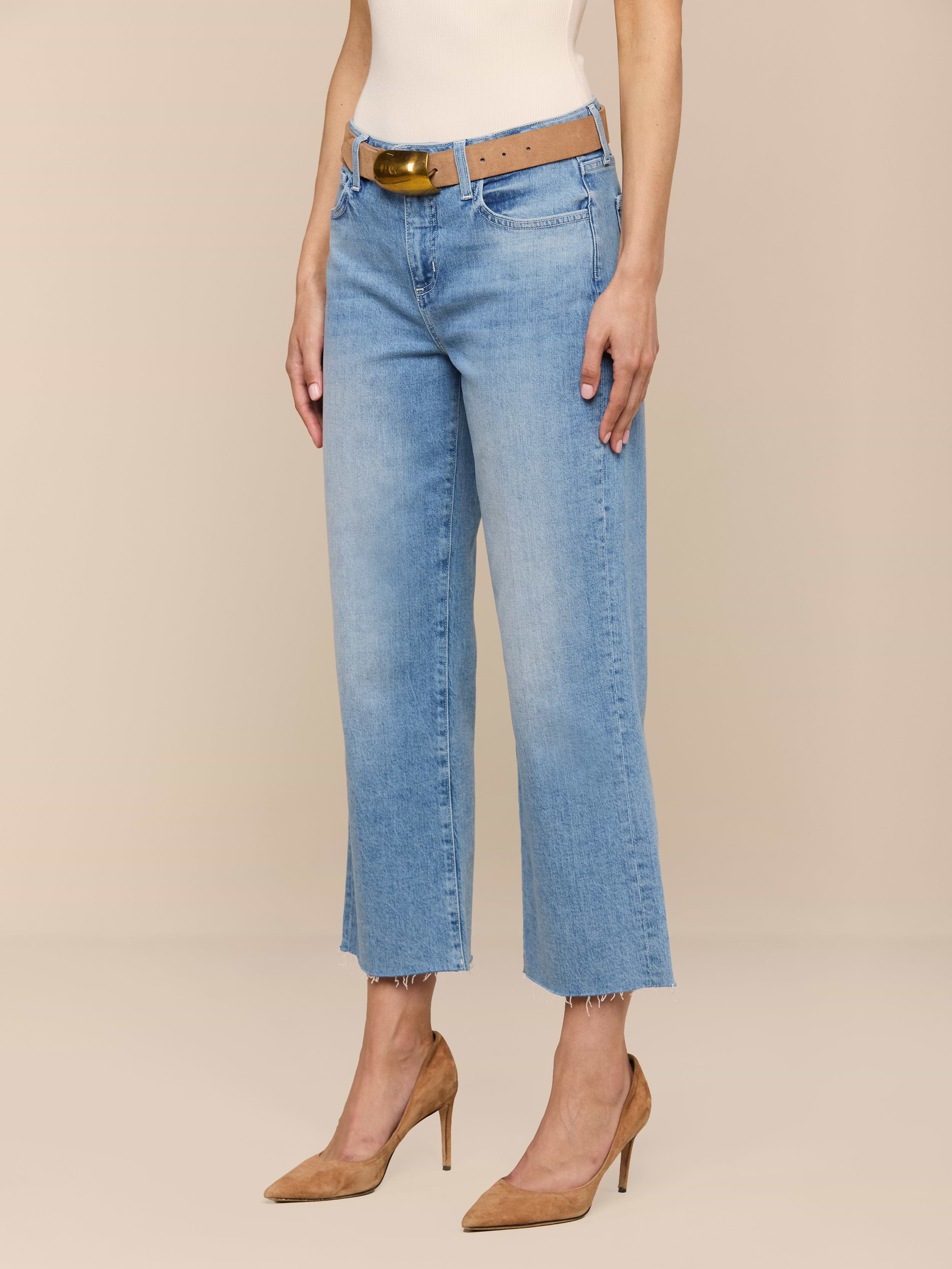 Eine Person modelliert die Wanda Cropped Wide-Leg Jean in hellblauer Waschung mit offenem Saum, gestylt mit einem hellbraunen Gürtel, einem weißen Oberteil und hellbraunen hochhackigen Schuhen vor einem schlichten beigen Hintergrund.