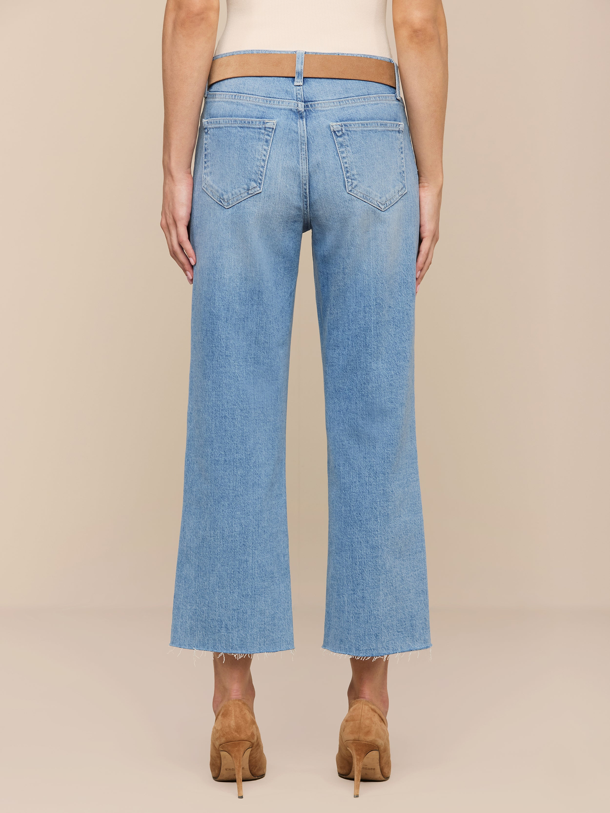 Eine Person ist von hinten in einer hellblauen Wanda Cropped Wide-Leg Jeans mit ausgefransten Säumen, hellbraunen Stöckelschuhen und einem ärmellosen beigen Oberteil vor einem neutralen Hintergrund abgebildet.