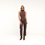 A woman wearing a sleeveless brown turtleneck, the Wanda Coated Cropped Wide-Leg Jean with a black belt, brown boots, and sunglasses stands against a beige background.