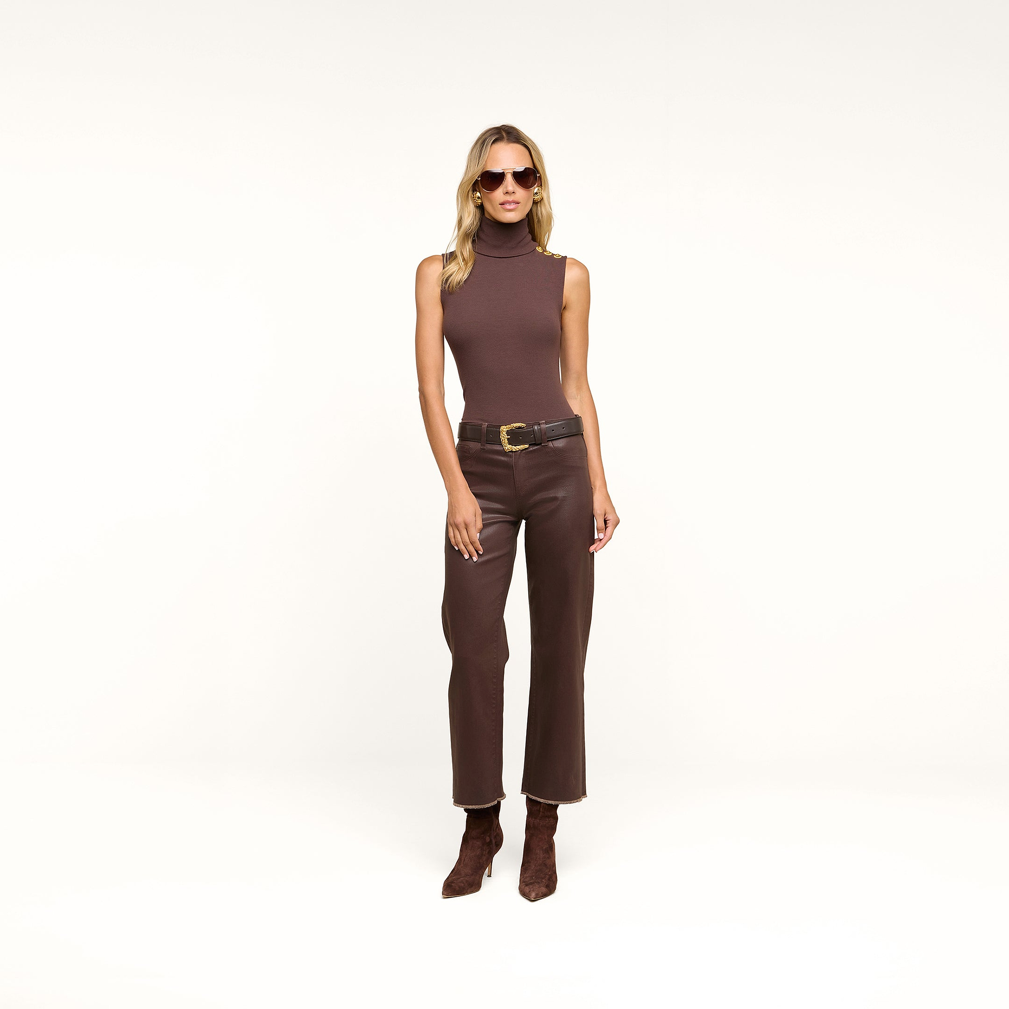 A woman wearing a sleeveless brown turtleneck, the Wanda Coated Cropped Wide-Leg Jean with a black belt, brown boots, and sunglasses stands against a beige background.