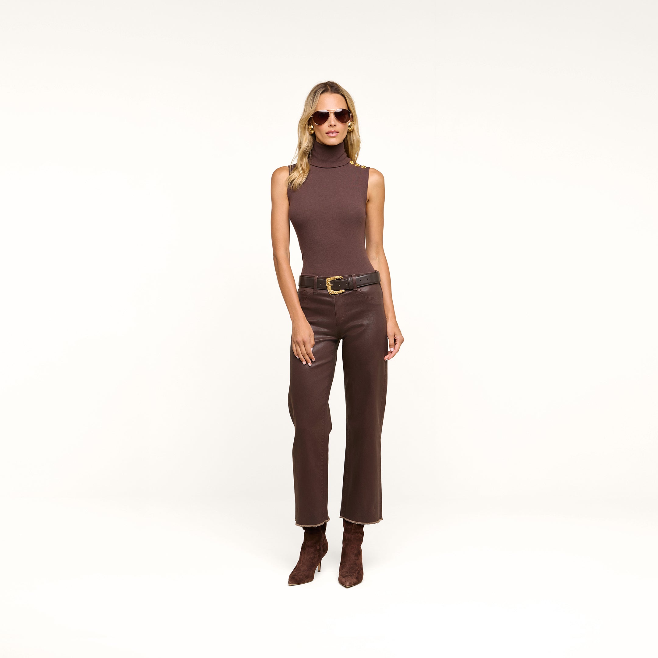 A woman wearing a sleeveless brown turtleneck, the Wanda Coated Cropped Wide-Leg Jean with a black belt, brown boots, and sunglasses stands against a beige background.