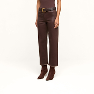 Wearing the Wanda Coated Cropped Wide-Leg Jean in brown, styled with a black belt featuring a gold buckle, a coordinating brown top, and pointed-toe boots, the individual channels the sleek look of coated denim while standing against a beige background.