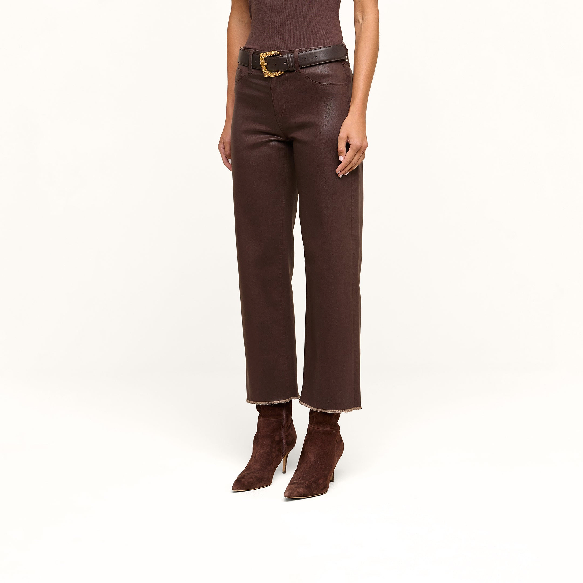 Wearing the Wanda Coated Cropped Wide-Leg Jean in brown, styled with a black belt featuring a gold buckle, a coordinating brown top, and pointed-toe boots, the individual channels the sleek look of coated denim while standing against a beige background.