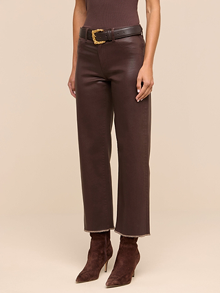 Wearing the Wanda Coated Cropped Wide-Leg Jean in brown, styled with a black belt featuring a gold buckle, a coordinating brown top, and pointed-toe boots, the individual channels the sleek look of coated denim while standing against a beige background.