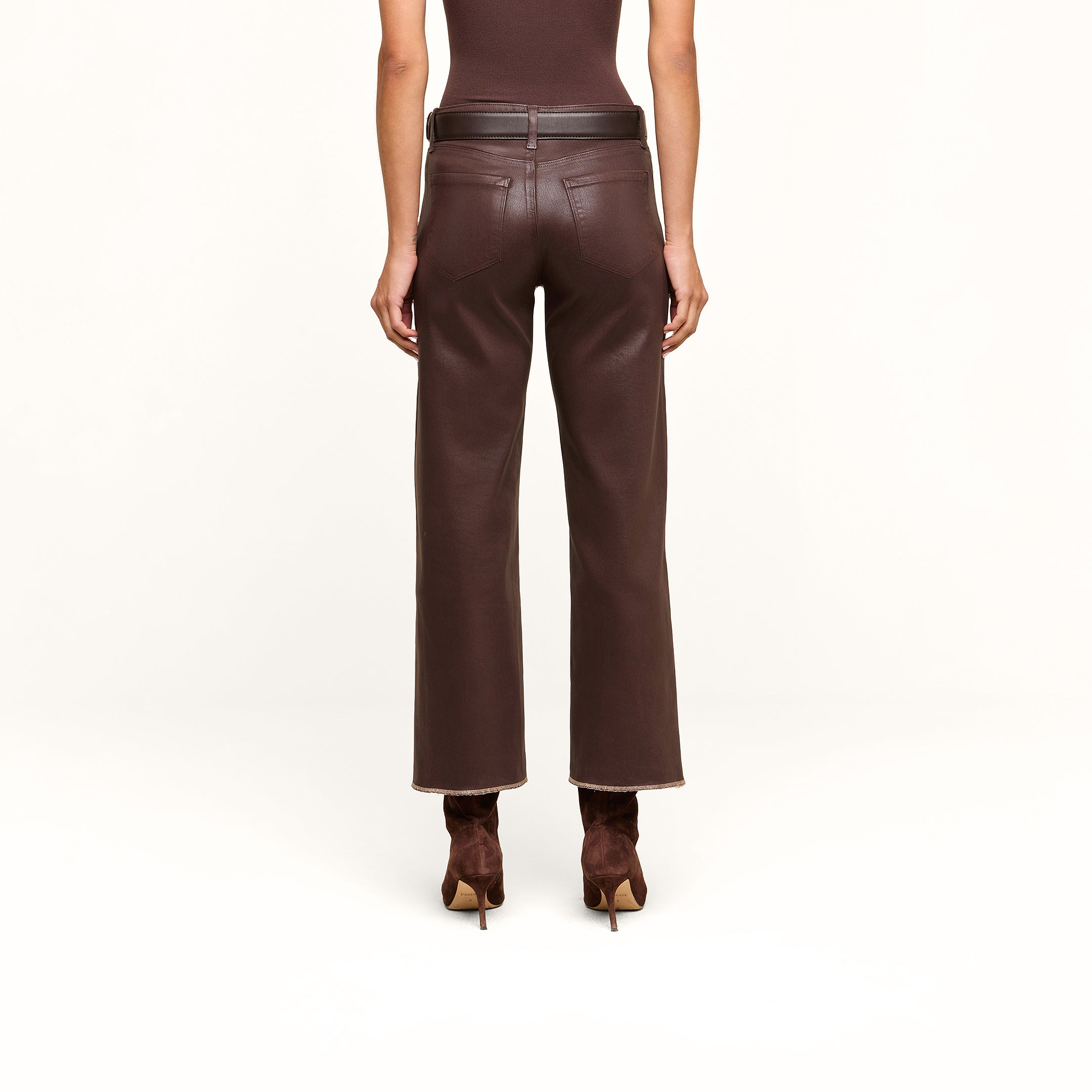A person stands facing away against a beige background, wearing the Wanda Coated Cropped Wide-Leg Jean—high-waisted brown coated denim pants with a raw hem—paired with a brown belt and matching brown heeled boots.