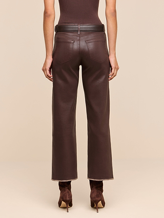 A person stands facing away against a beige background, wearing the Wanda Coated Cropped Wide-Leg Jean—high-waisted brown coated denim pants with a raw hem—paired with a brown belt and matching brown heeled boots.