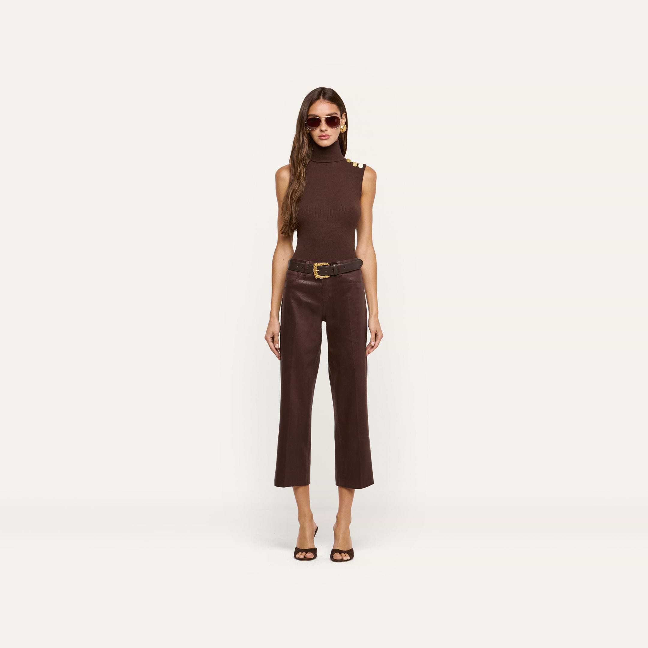 A woman stands against a plain white background wearing a sleeveless dark java brown top with gold buttons, the Wanda Coated Cropped Wide-Leg Jean in brown, a black belt with a gold buckle, sunglasses, and heeled sandals.