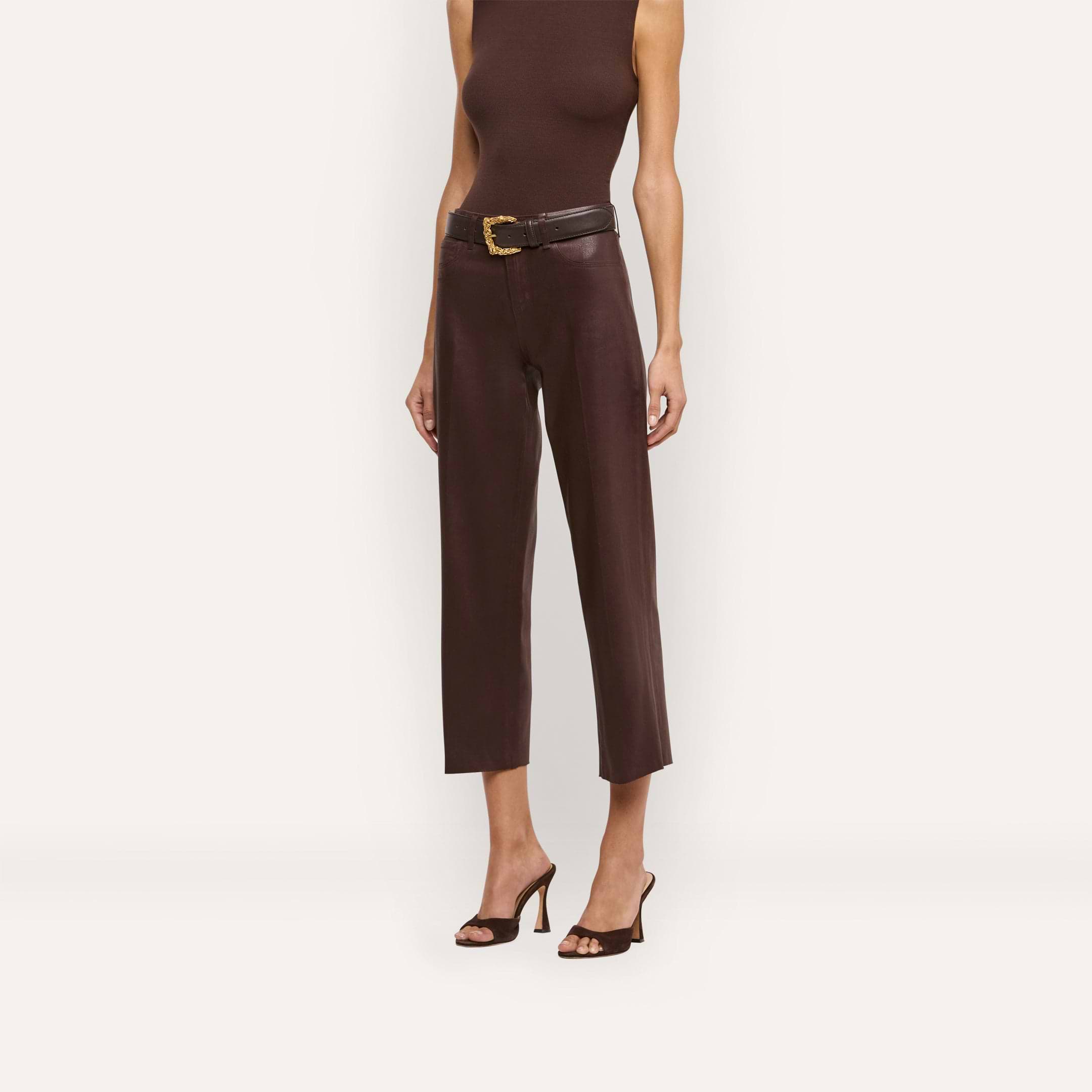 Wearing a sleeveless brown top and the Wanda Coated Cropped Wide-Leg Jean in dark java brown, styled with a belt and black heeled sandals, a person stands against a plain light background.
