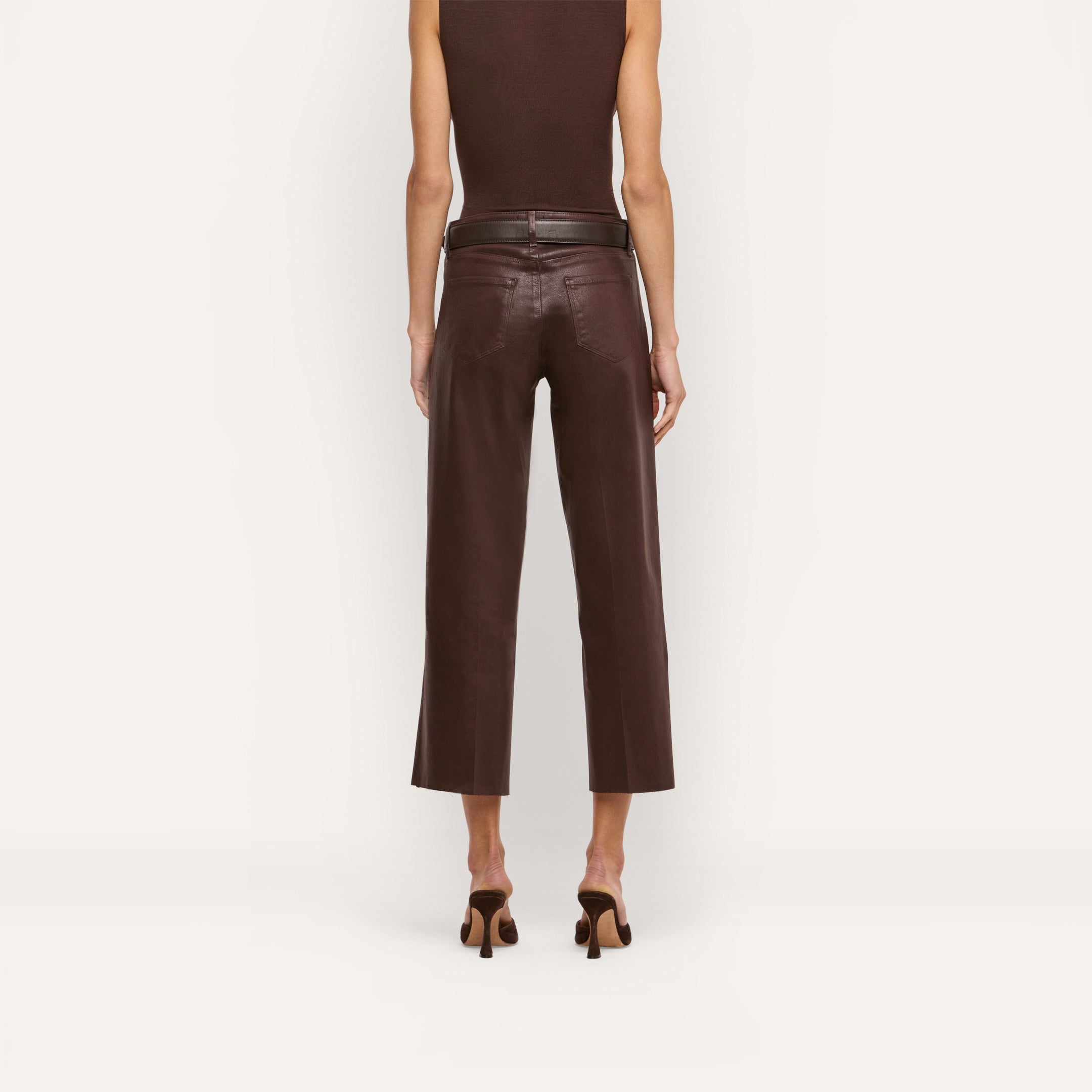 A person stands facing away against a plain background, wearing the Wanda Coated Cropped Wide-Leg Jean in dark java brown, paired with a sleeveless top and heeled sandals.