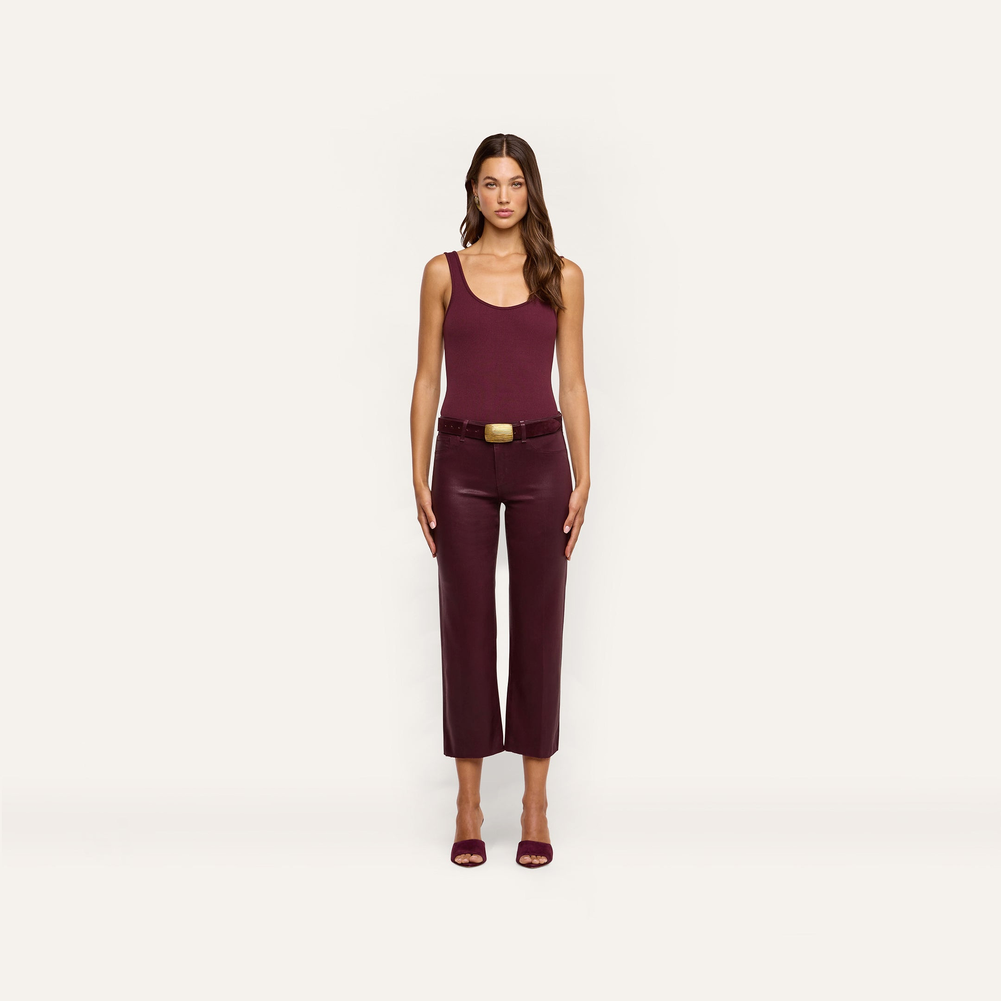 A woman stands facing forward, wearing a sleeveless burgundy top, the Wanda Coated Cropped Wide-Leg Jean with a belt, and burgundy heels against a plain light background.