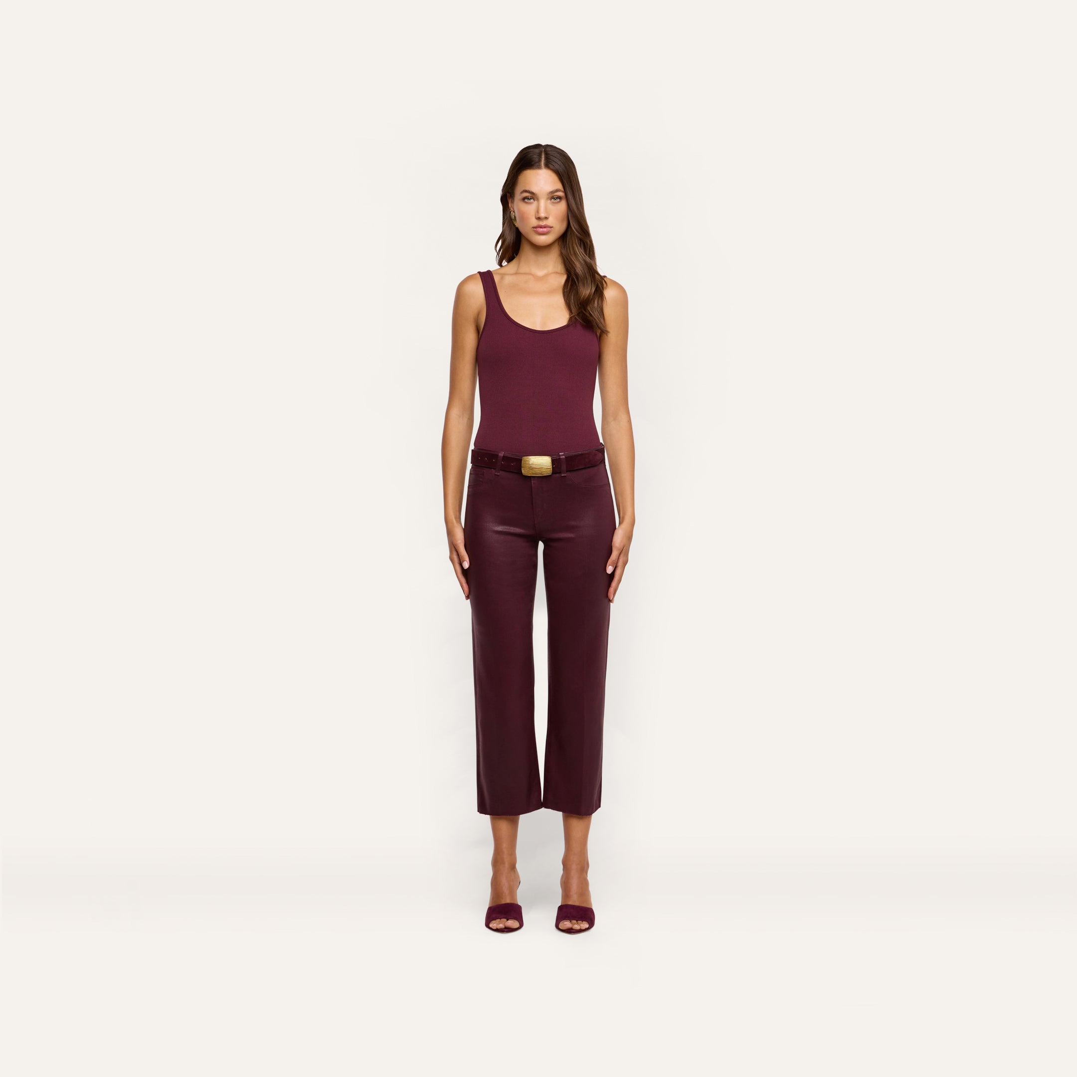 A woman stands facing forward, wearing a sleeveless burgundy top, the Wanda Coated Cropped Wide-Leg Jean with a belt, and burgundy heels against a plain light background.