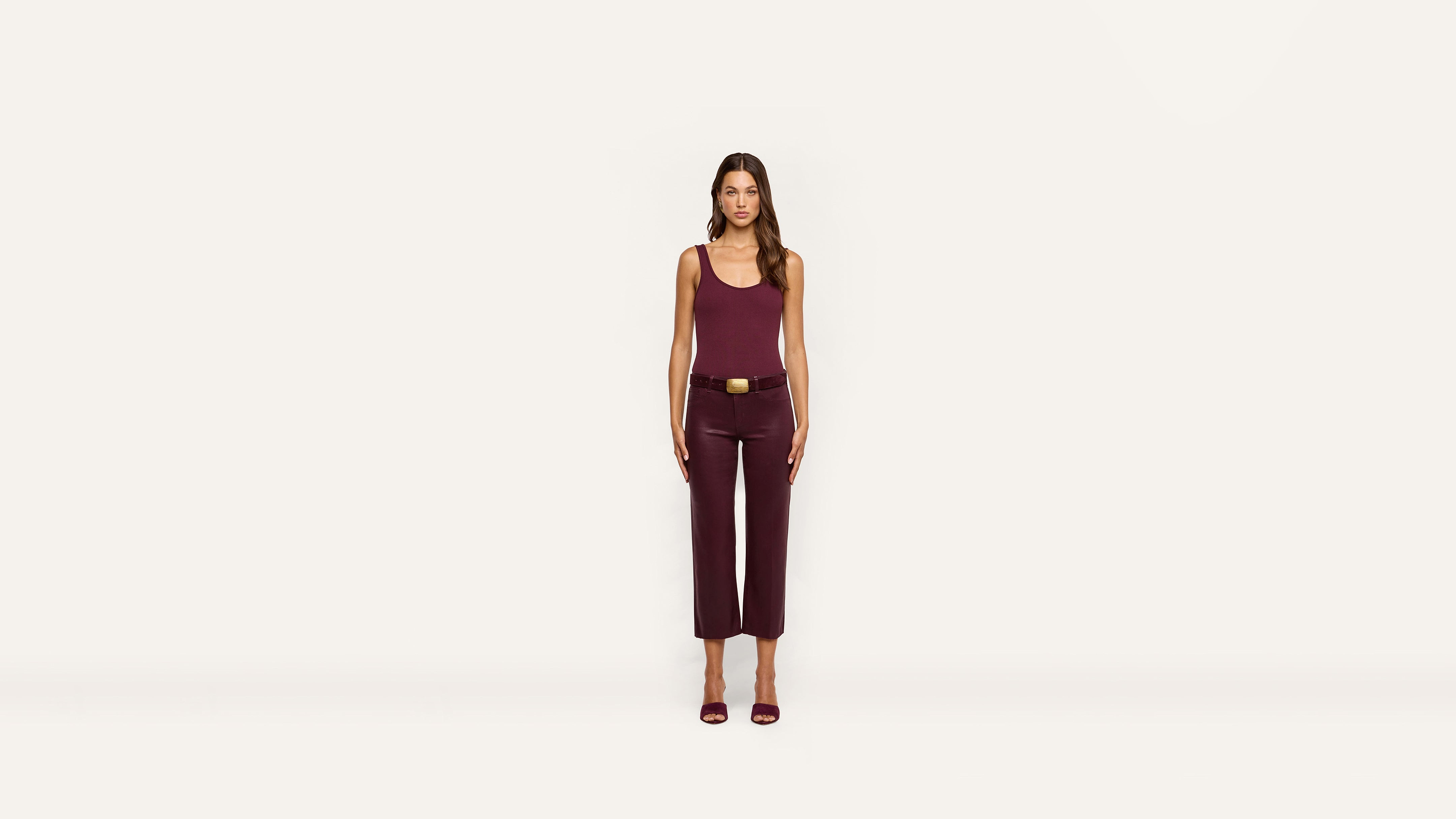 A woman stands facing forward against a plain white background, dressed in a sleeveless maroon top, matching cropped pants, a black belt, and maroon heeled sandals.