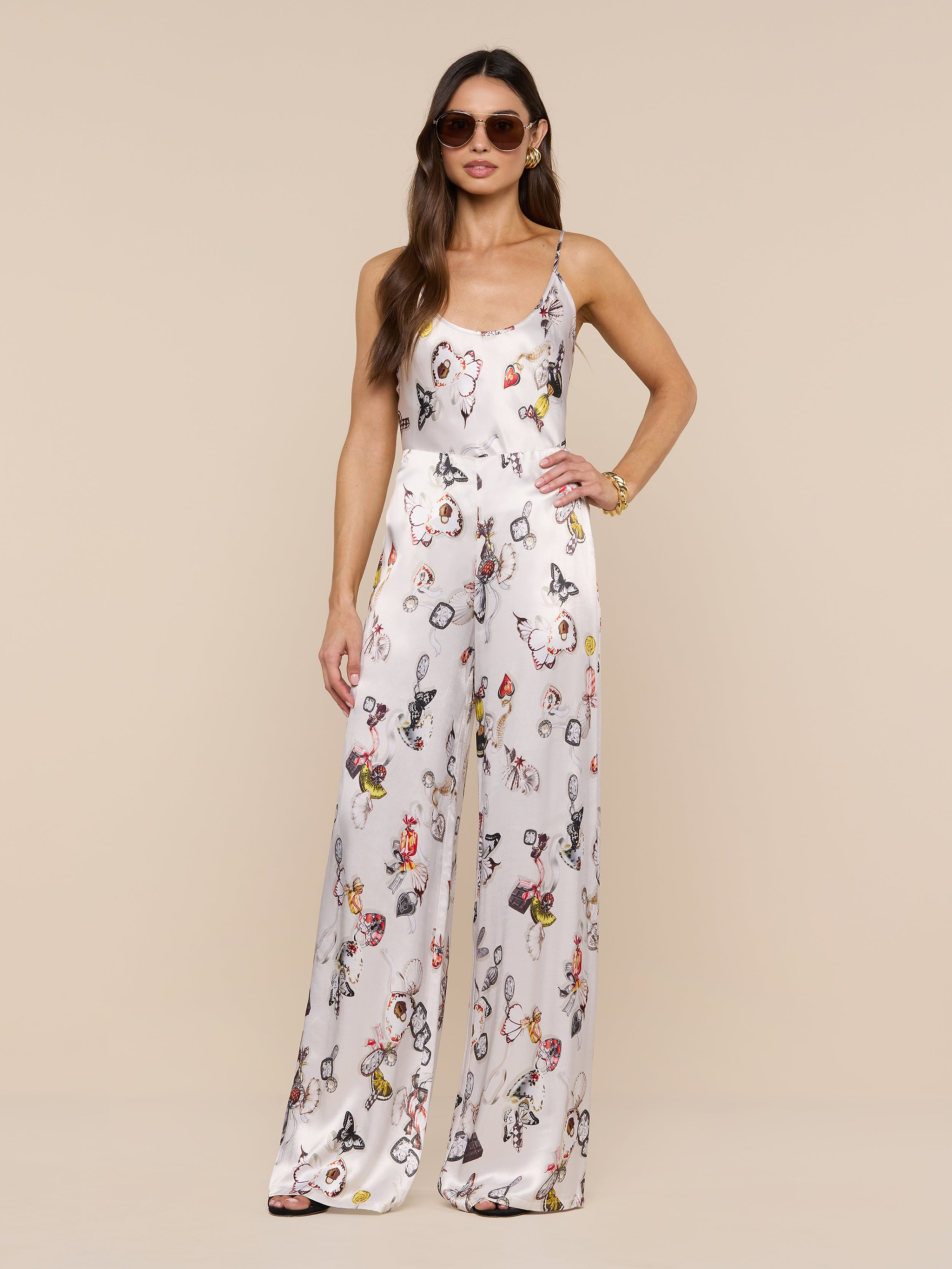 A woman in sunglasses stands confidently against a plain white background, wearing the Kimia Satin Pant—a white, sleeveless jumpsuit featuring a colorful butterfly print and straight-leg pants.