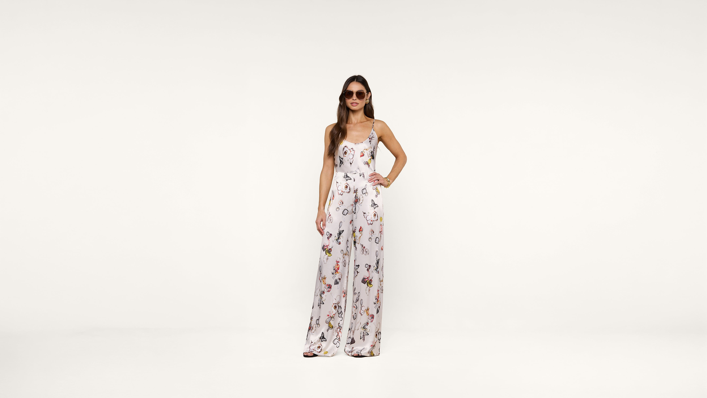 A woman wearing sunglasses and a white floral jumpsuit stands confidently against a plain white background with one hand on her hip.