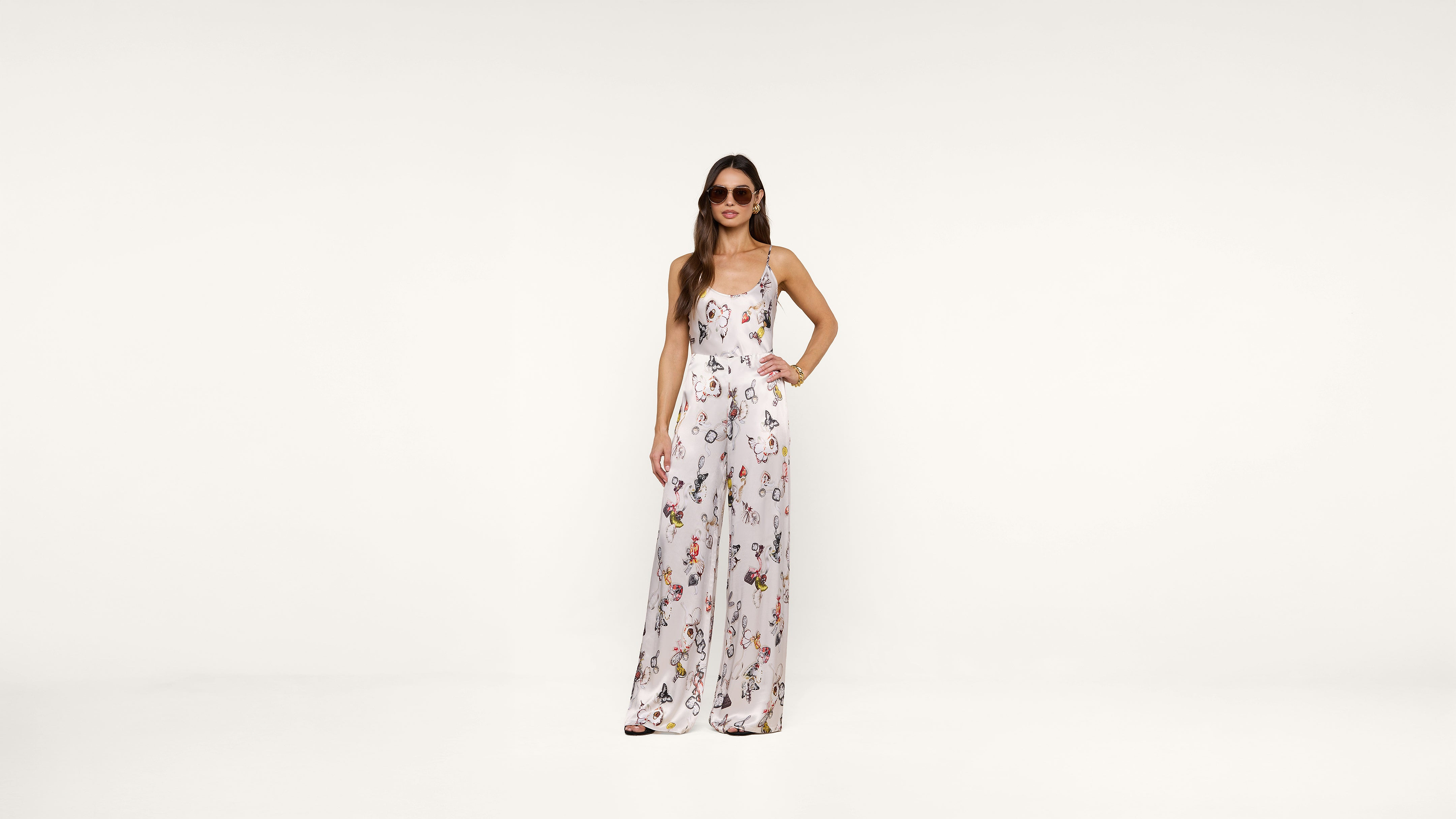 A woman wearing sunglasses and a white floral jumpsuit stands confidently against a plain white background with one hand on her hip.