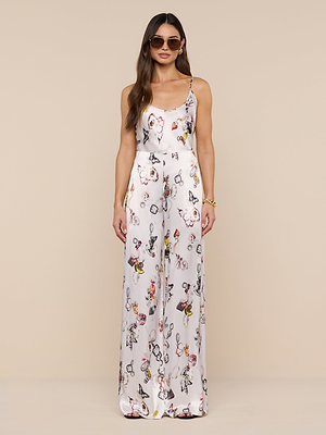 A woman wearing sunglasses and a white, sleeveless jumpsuit with a colorful abstract print and high-rise, straight-leg Kimia Satin Pant stands facing forward against a plain beige background.