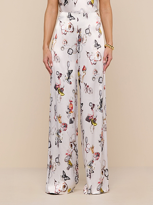 A person wears the Kimia Satin Pant—high-rise, wide-leg white satin pants with a colorful whimsical print of illustrated characters and objects—standing against a neutral background.