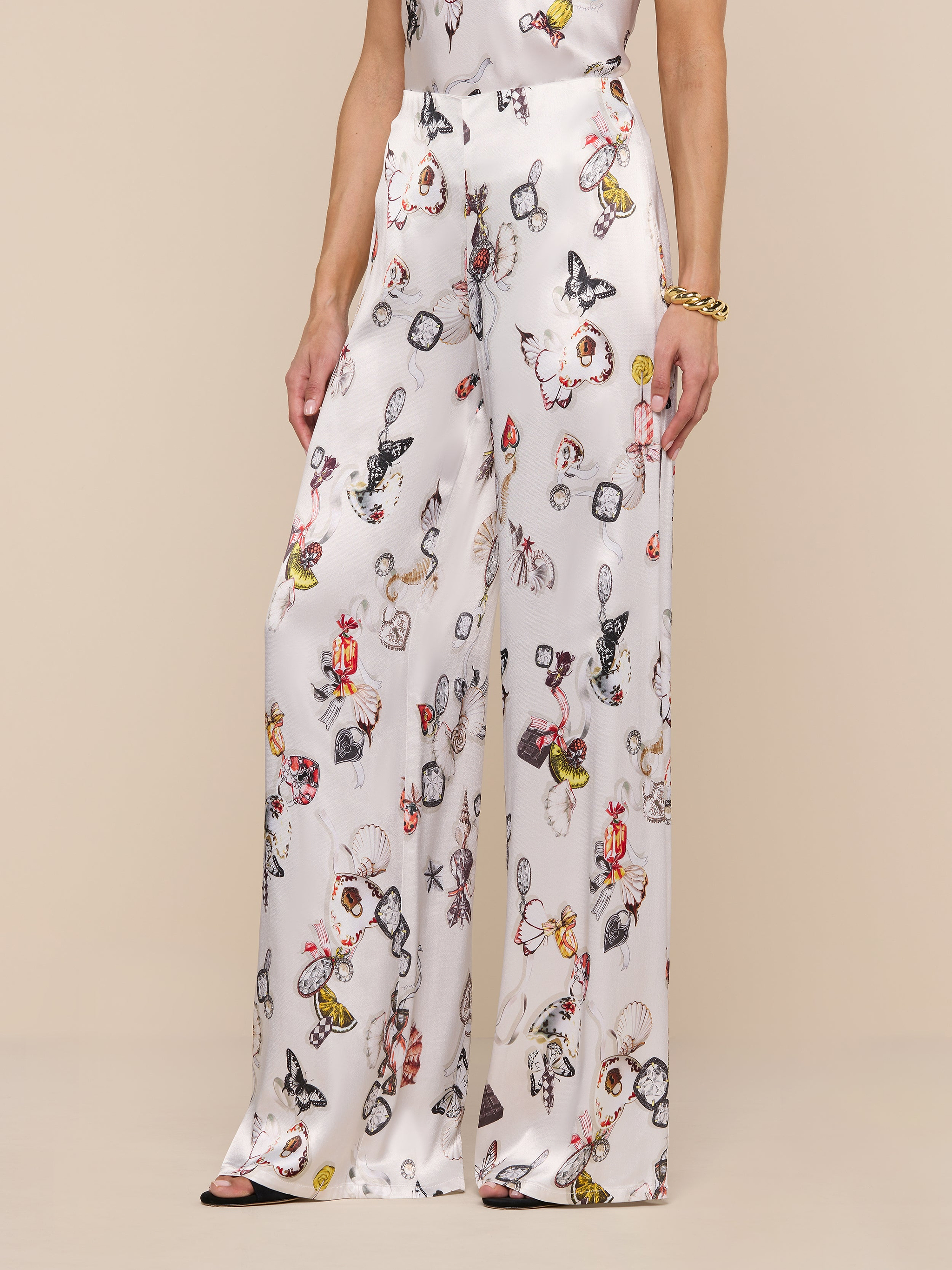 A person wearing the Kimia Satin Pant—a high-rise, silky white satin wide-leg pant featuring a colorful butterfly print—stands against a plain white background. Only the lower half of the body is visible.