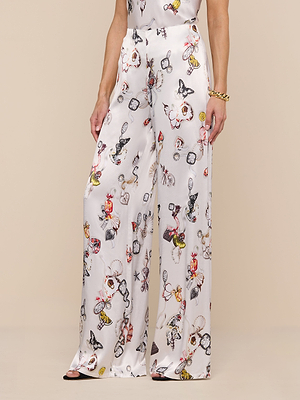 Wearing the Kimia Satin Pant—high rise, wide-leg satin pants with a white base and playful, colorful print of illustrated faces and objects—paired with a gold bracelet and sleek black heels.