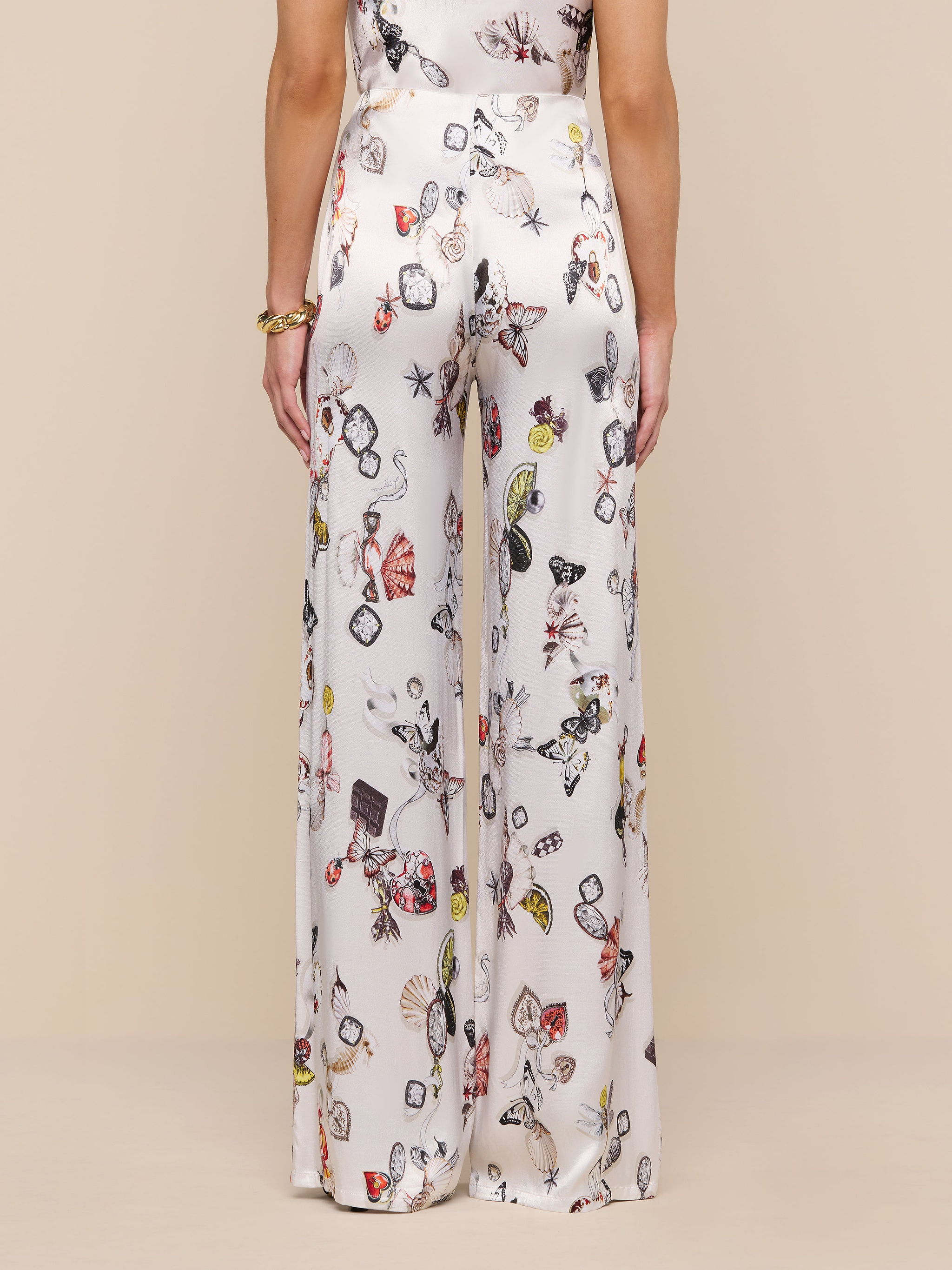A woman is shown from the back wearing the Kimia Satin Pant—high-rise, wide-leg white pants featuring a colorful jewelry print—standing against a plain white background.