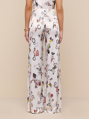 A person is seen from the back wearing the Kimia Satin Pant, a high rise, white satin straight-leg style with a colorful butterfly print, standing against a beige background.