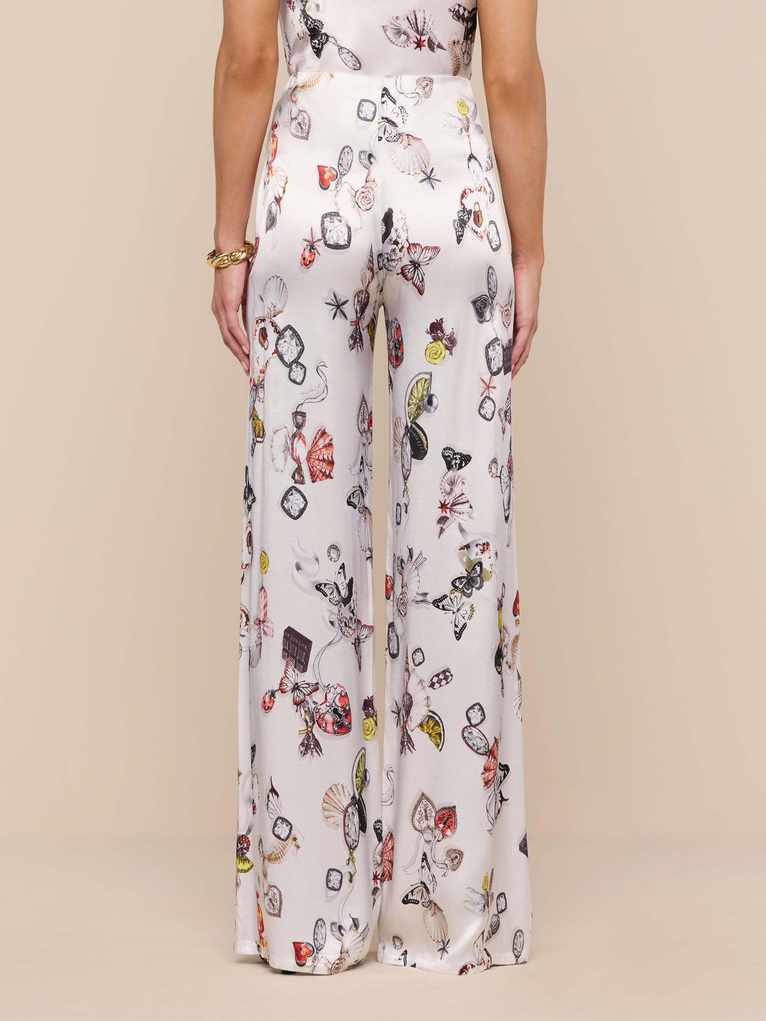 A woman is shown from the back wearing the Kimia Satin Pant—high-rise, wide-leg white pants featuring a colorful jewelry print—standing against a plain white background.