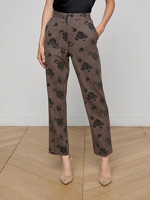 A person wears the L'AGENCE Rebel Flocked Trouser—high-rise brown straight-leg trousers with a black floral pattern—paired with a black top and beige pointed-toe heels, standing on a wooden floor with hands in pockets.