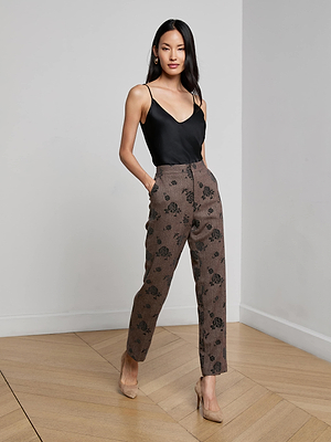 A woman poses indoors against a plain white wall wearing L'AGENCE Rebel Flocked Trouser with a black floral pattern, paired with a black sleeveless V-neck top and nude heels. She has one hand in her pocket.