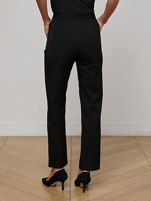 A person in Rebel Trouser high-rise, straight-leg dress pants and black heels stands on a wooden floor, back to camera, with hands in pockets.