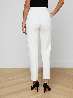 A woman stands on a wooden floor, facing away from the camera, wearing white Rebel Trousers in stretch suit fabric, a black sleeveless top, and black high-heeled shoes against a plain white wall.