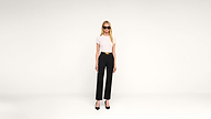 Woman with long blonde hair wearing black sunglasses, a white short-sleeve top, black high-waisted pants with a belt, and black heels stands against a plain white background.