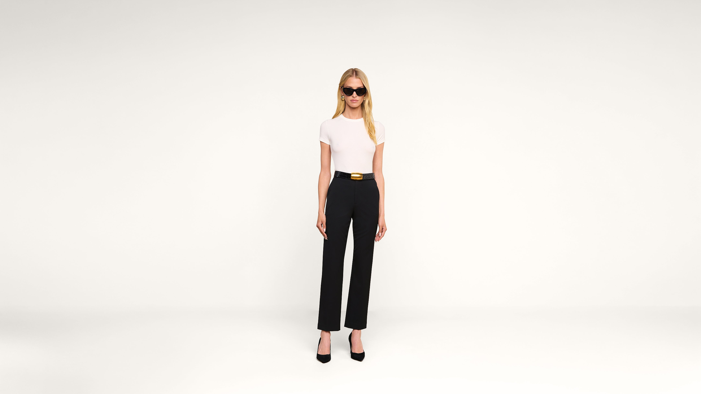 Woman with long blonde hair wearing black sunglasses, a white short-sleeve top, black high-waisted pants with a belt, and black heels stands against a plain white background.