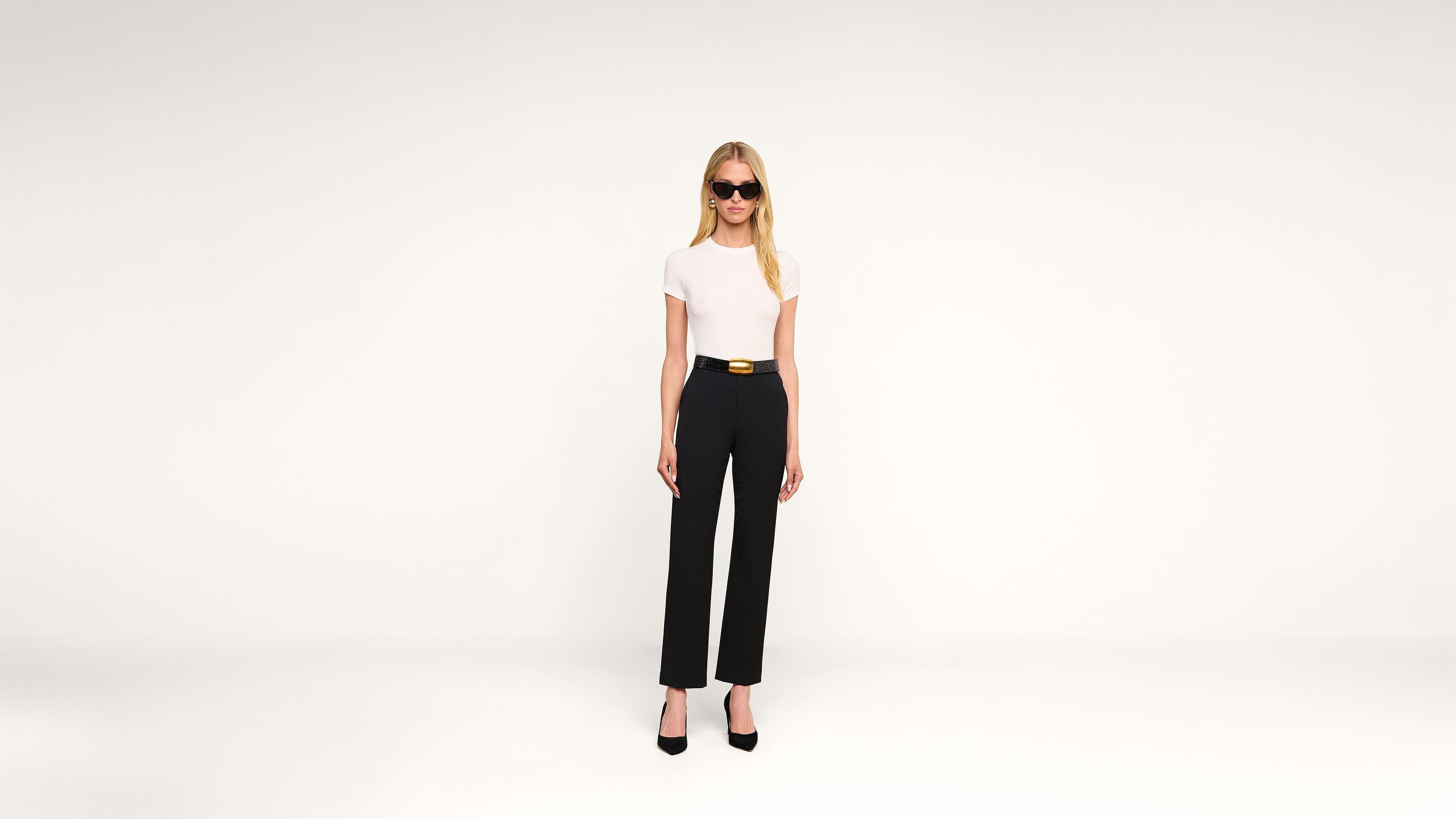 Woman with long blonde hair wearing black sunglasses, a white short-sleeve top, black high-waisted pants with a belt, and black heels stands against a plain white background.