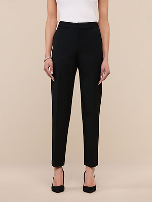 A woman stands against a plain background wearing the Rebel Trouser—black Italian stretch suiting, high-rise, straight-leg—paired with a white sleeveless top, black pointed-toe heels, and a bracelet on her right wrist. Her arms are relaxed at her sides.