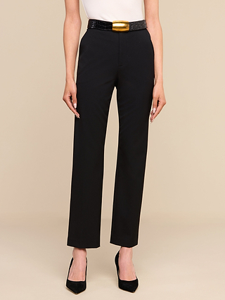 تقف امرأة ترتدي بنطال «Rebel Trouser» — وهو بنطال أسود بخصر مرتفع وساق مستقيمة، مصنوع من قماش إيطالي مرن مخصص للبدلات — مع بلوزة بيضاء وحزام أسود مزود بمشبك ذهبي كبير، وحذاء كعب عالٍ أسود ذي مقدمة مدببة، أمام خلفية باللون البيج.