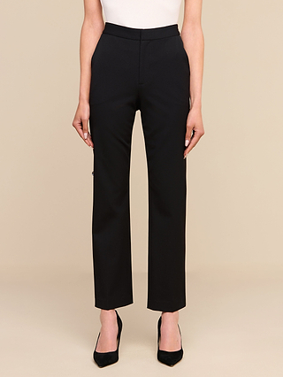 تقف امرأة ترتدي بنطال «Rebel Trouser» باللون الأسود — وهو بنطال بخصر مرتفع وساق مستقيمة مصنوع من قماش بدلات إيطالي مرن — مع حذاء بكعب عالٍ أسود، أمام خلفية بيج سادة، وذراعاها مسترخيتان على جانبي جسدها.
