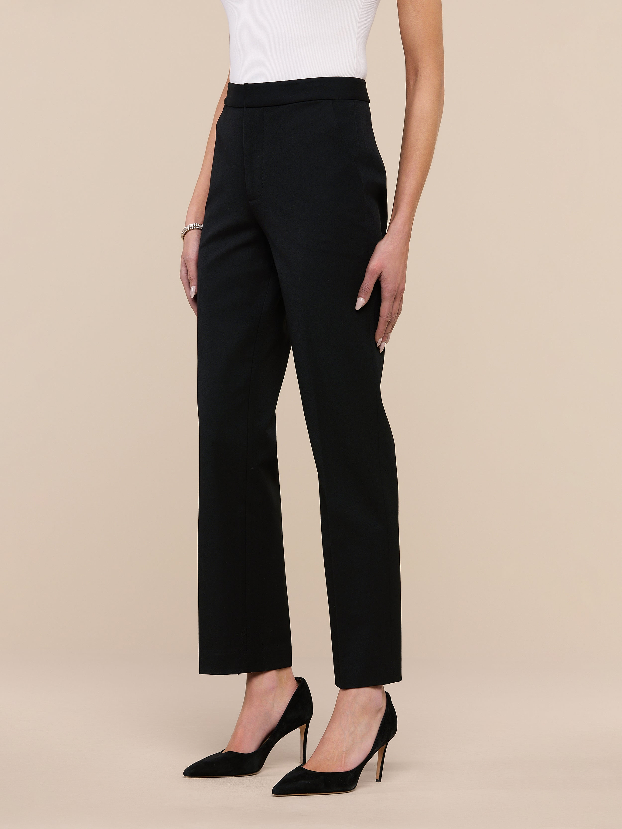 A person wearing the Rebel Trouser—high-rise, straight-leg black trousers crafted from Italian stretch suiting—along with a white top, black heels, and a black belt with a gold buckle stands against a plain light background.