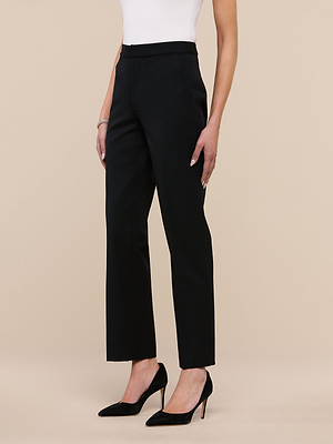 A person stands against a plain beige background wearing high-rise black Rebel Trousers, a white sleeveless top, and black high-heeled shoes.