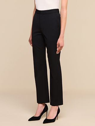 تقف امرأة ترتدي بنطال «Rebel Trouser» — وهو بنطال بخصر مرتفع وساق مستقيمة مصنوع من قماش إيطالي مرن — مع بلوزة بيضاء وحذاء أسود بكعب عالٍ، أمام خلفية بيج. ولا يظهر سوى النصف السفلي من جسدها.