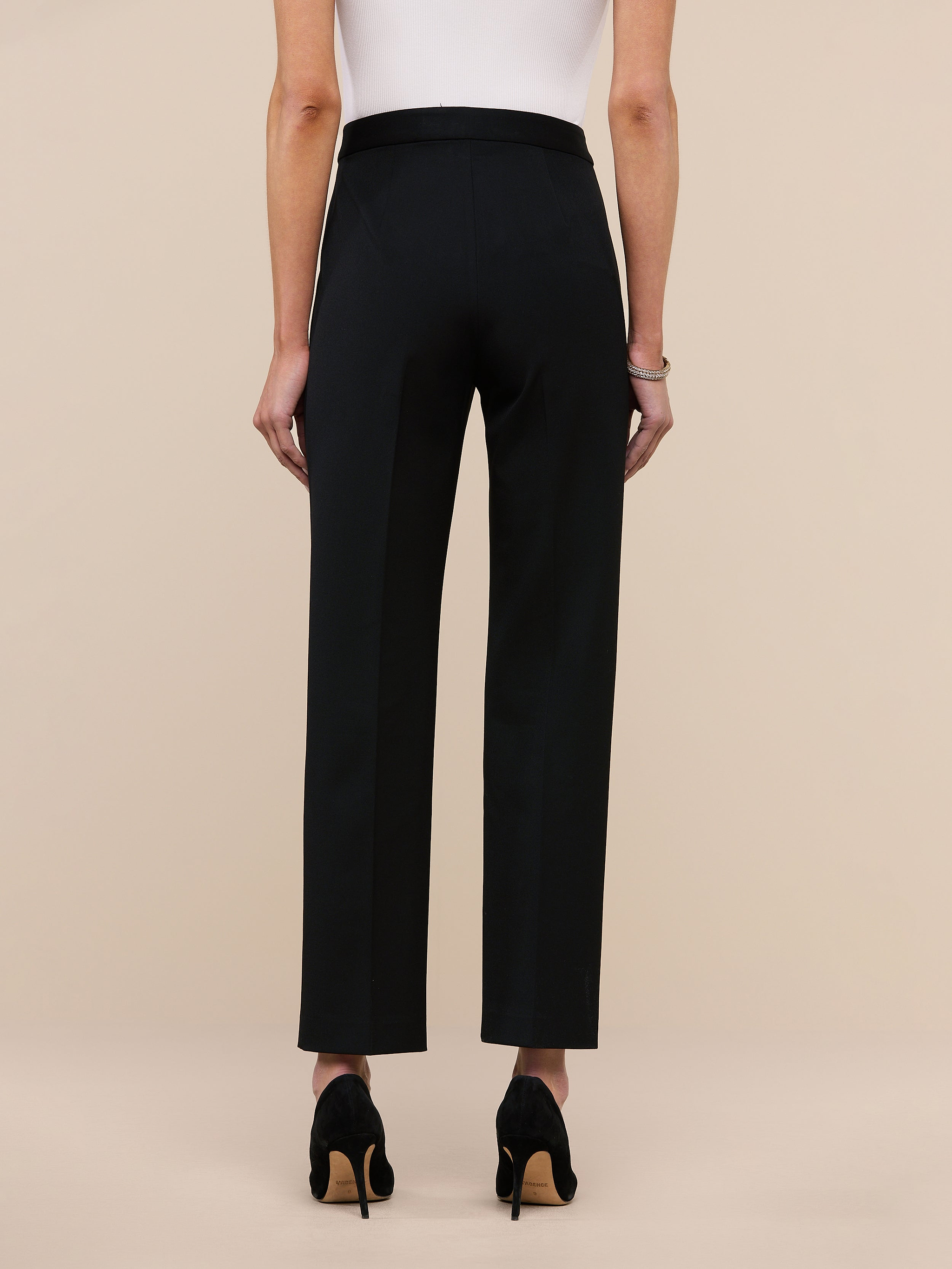 A person, seen from behind against a plain white background, is wearing the Rebel Trouser—black high-rise straight-leg pants crafted from Italian stretch suiting—paired with black high heels and a sleeveless light-colored top.