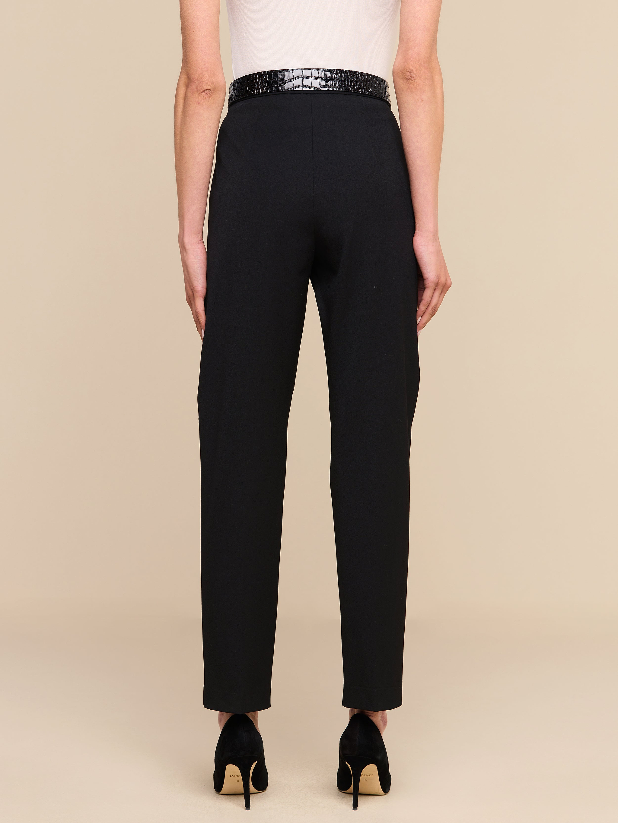 Rückansicht einer Person, die die „Rebel Trouser“ trägt – eine schwarze, hoch taillierte italienische Stretch-Anzughose mit geradem Bein –, kombiniert mit einem strukturierten schwarzen Gürtel, schwarzen Pumps und einem weißen, ärmellosen Oberteil; die Person steht vor einem beigen Hintergrund.