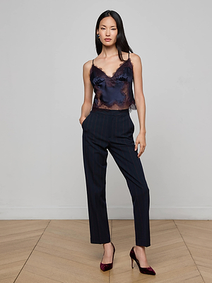 A woman with long dark hair stands against a light gray wall, hands in her pockets, wearing L'AGENCE Rebel Pinstriped Trouser, a navy lace-trimmed camisole, and burgundy heels.