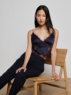 A woman with long black hair sits on a wooden chair, wearing the L'AGENCE Kimber Silk Lace Camisole and dark pinstriped pants, gazing confidently at the camera against a plain, light background.