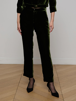 A person stands on a light wood floor wearing L'AGENCE Rebel Velvet Trousers in olive with a matching belt, paired with sheer black socks and black pointed-toe heels. Their hands are by their sides, and their upper body is mostly out of frame.