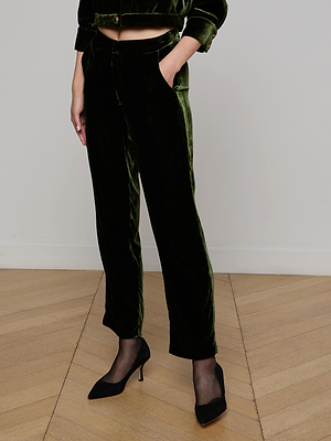 Indoors, a person wears L'AGENCE Rebel Velvet Trouser in olive with a matching jacket, sheer black tights, and black heels; their hands are in their pockets, and their head is out of frame.