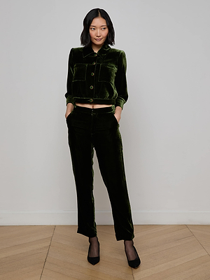 A woman with shoulder-length dark hair stands indoors against a white wall, wearing L'AGENCE Rebel Velvet Trousers and a matching olive button-up jacket with black heels, hands in her pockets.